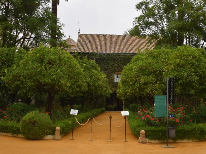 Jardines del palacio de las dueñas, un cartel de audioguia y otro indicando el recorrido, casita con balcón, plantas, árboles y flores